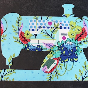 Może przedstawiać: Jasnoniebieski wykrój z tkaniny w kształcie zabytkowej maszyny do szycia. Maszyna ozdobiona jest wzorami kwiatowymi i liściastymi w odcieniach niebieskiego, zielonego, czerwonego i żółtego. Na maszynie widoczne jest różowe serce.