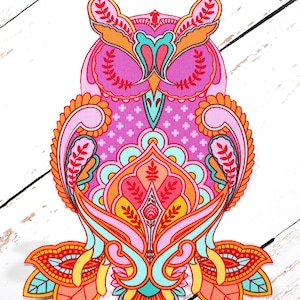 Może przedstawiać: Kolorowa aplikacja w kształcie sowy ze skomplikowanymi wzorami paisley i kwiatowymi. Projekt zawiera odcienie różu, pomarańczy, żółci i turkusu. Aplikacja jest umieszczona na białym drewnianym tle.