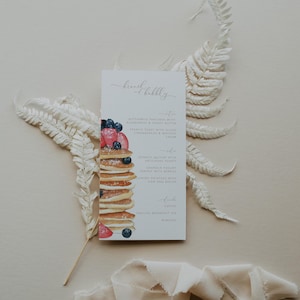 May include: A white menu card with the text "brunch & bubbly" at the top. The menu lists breakfast items such as buttermilk pancakes with blueberries and honey butter, spinach quiche with arugula, and granola yogurt. An illustration of a stack of pancakes with berries and syrup is shown on the left side of the card.