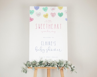 May include: A white sign with a pink and blue border announcing a baby shower for Claire. The sign is decorated with colorful candy hearts with messages like "Sweetheart" and "All Mine". The text on the sign reads "little sweetheart is on her way. Welcome to Claire's Baby Shower. Aug 15 20xx Dallas."