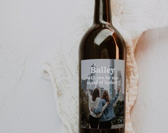 Op de afbeelding: Een donkere glazen wijnfles met een aangepast rechthoekig etiket. Het etiket toont een foto van twee personen die elkaar omhelzen voor een skyline, met de tekst Bailey, will you be my maid of honor?