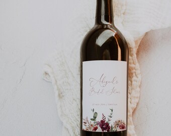 Puede incluir: Una botella de vino oscuro con una etiqueta blanca con el texto "Abigail's Bridal Shower" en escritura elegante. La etiqueta está adornada con ilustraciones florales en tonos burdeos y verdes. La botella está sobre una tela blanca.