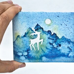 May include: A blue and green watercolor painting of a mountain range with a white silhouette of a deer in the foreground. The painting has a white moon in the upper right corner.