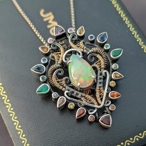 May include: A handcrafted pendant necklace featuring a large, pear-shaped opal as the centerpiece. The opal is surrounded by a complex wire-wrapped design with silver and gold-toned metal, accented by various colorful gemstones. The necklace rests on a black box.