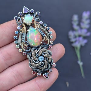 May include: A handcrafted pendant featuring an opal gemstone with a pearlescent sheen, accented by smaller gemstones in various colors. The pendant has intricate wirework in silver and gold tones, with a floral-like design. The overall aesthetic is artistic and unique.