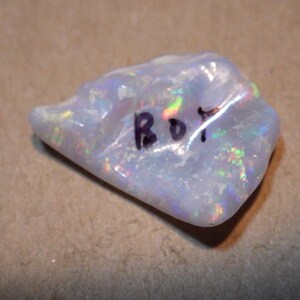 May include: A white opal gemstone with iridescent rainbow colors. The stone is rough and has a natural shape. It is labeled "BOT" in black ink.