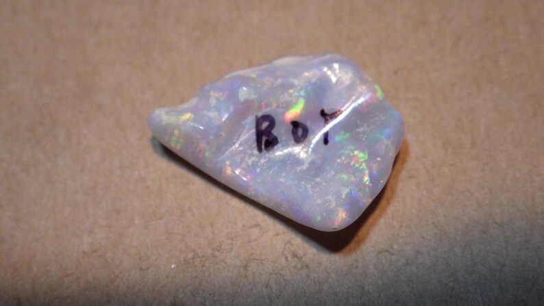 May include: A white opal gemstone with iridescent rainbow colors. The stone is rough and has a natural shape. It is labeled "BOT" in black ink.