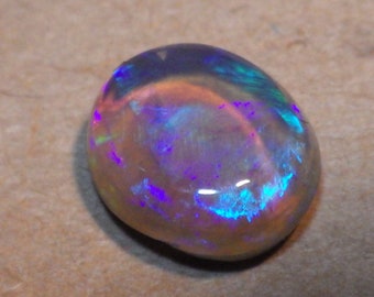 May include: A round, oval-shaped opal gemstone with a light gray base and iridescent blue, purple, and green flashes.