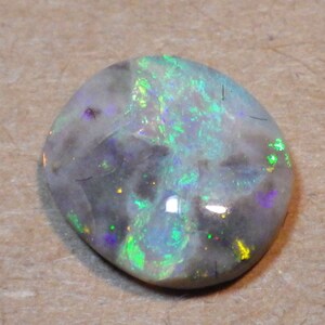 May include: A white opal cabochon with a green, blue, and purple iridescent sheen. The opal has a gray and black matrix.