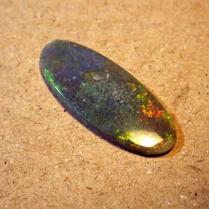 May include: A green, blue, and orange opal cabochon with a smooth, polished surface. The opal is oval-shaped and has a light, iridescent sheen.