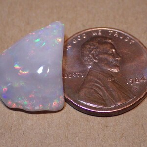 May include: A white opal gemstone with iridescent rainbow flashes, next to a US penny for size comparison.