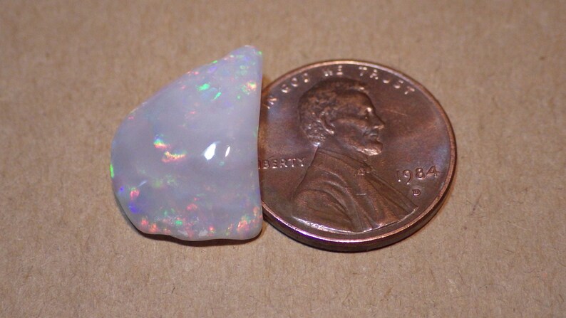 May include: A white opal gemstone with iridescent rainbow flashes, next to a US penny for size comparison.