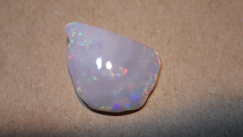May include: A white opal gemstone with iridescent flashes of color. The opal is irregularly shaped and has a smooth, polished surface.