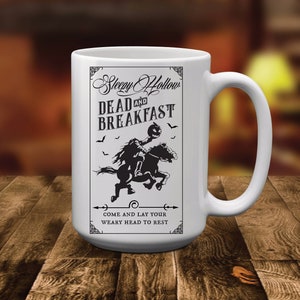 Op de afbeelding: Een witte keramische mok met een zwarte afbeelding van een ruiter zonder hoofd die een pompoen vasthoudt terwijl hij op een paard rijdt. De tekst luidt Sleepy Hollow DEAD AND BREAKFAST en COME AND LAY YOUR WEARY HEAD TO REST, met decoratieve hoeken.