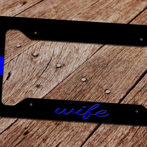 May include: Black license plate frame with the word "wife" in blue script. The frame has a blue stripe on the left and right sides. The background is a wooden surface.
