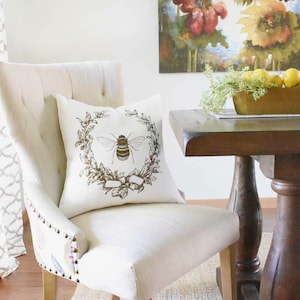 May include: A white chair with a beige cushion and a decorative pillow featuring a brown bee in a wreath. The pillow has a white background with a brown bee in a wreath of leaves and a brown ribbon.