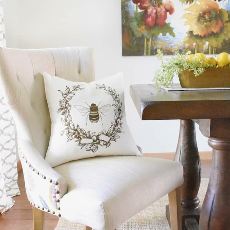 May include: A white chair with a beige cushion and a decorative pillow featuring a brown bee in a wreath. The pillow has a white background with a brown bee in a wreath of leaves and a brown ribbon.