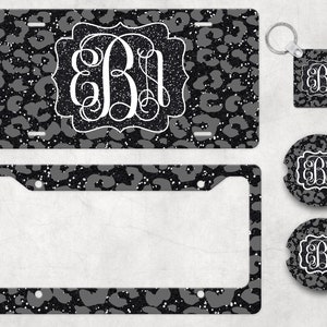 May include: A set of black and white leopard print car accessories featuring a monogrammed initial. The set includes a license plate frame, a keychain, a coaster, and a square sticker.