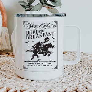 Op de afbeelding: Een witte roestvrijstalen reismok met een handvat en een zwarte deksel. De voorzijde bevat een zwarte afbeelding van een ruiter zonder hoofd op een paard, met de tekst Sleepy Hollow DEAD AND BREAKFAST en COME AND LAY YOUR WEARY HEAD TO REST.