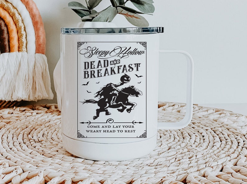 Op de afbeelding: Een witte roestvrijstalen reismok met een handvat en een zwarte deksel. De voorzijde bevat een zwarte afbeelding van een ruiter zonder hoofd op een paard, met de tekst Sleepy Hollow DEAD AND BREAKFAST en COME AND LAY YOUR WEARY HEAD TO REST.