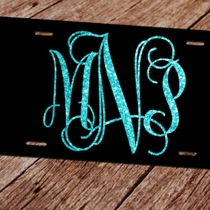 Puede incluir: Una placa de matr&iacute;cula negra con un monograma de purpurina turquesa de las letras "M" y "S" en una fuente cursiva.