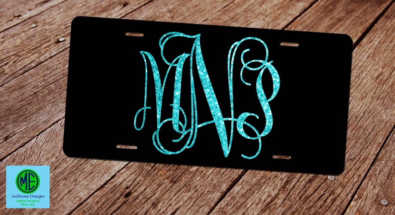 Puede incluir: Una placa de matr&iacute;cula negra con un monograma de purpurina turquesa de las letras "M" y "S" en una fuente cursiva.