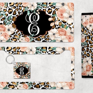 May include: A set of car accessories featuring a leopard print with floral accents. The set includes a license plate frame, a key chain, and a seatbelt cover. The license plate frame and seatbelt cover have a black border. The key chain has a small charm with a black background and a white letter "E" in the center.