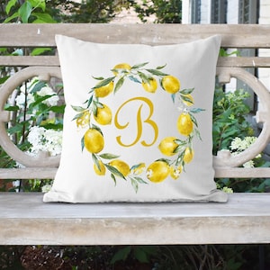 May include: White throw pillow with a lemon wreath design and the letter "B" in the center. The wreath is made of yellow lemons and green leaves.