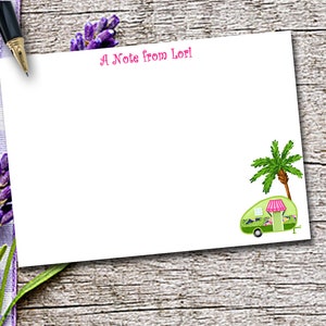 May include: A white rectangular stationery card featuring the pink text A Note from Lori at the top and a green camper with a pink striped awning and a palm tree in the bottom right corner. The card is positioned next to a black fountain pen.