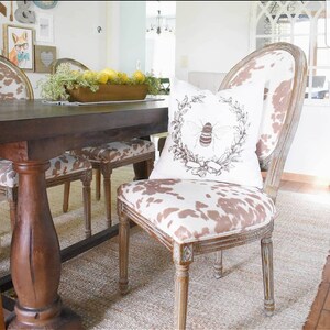 May include: A wooden dining table with a brown top and a single pedestal base.  The table is set with a bowl of lemons and greenery.  Two chairs with brown and white cowhide upholstery and a bee-themed pillow are visible.