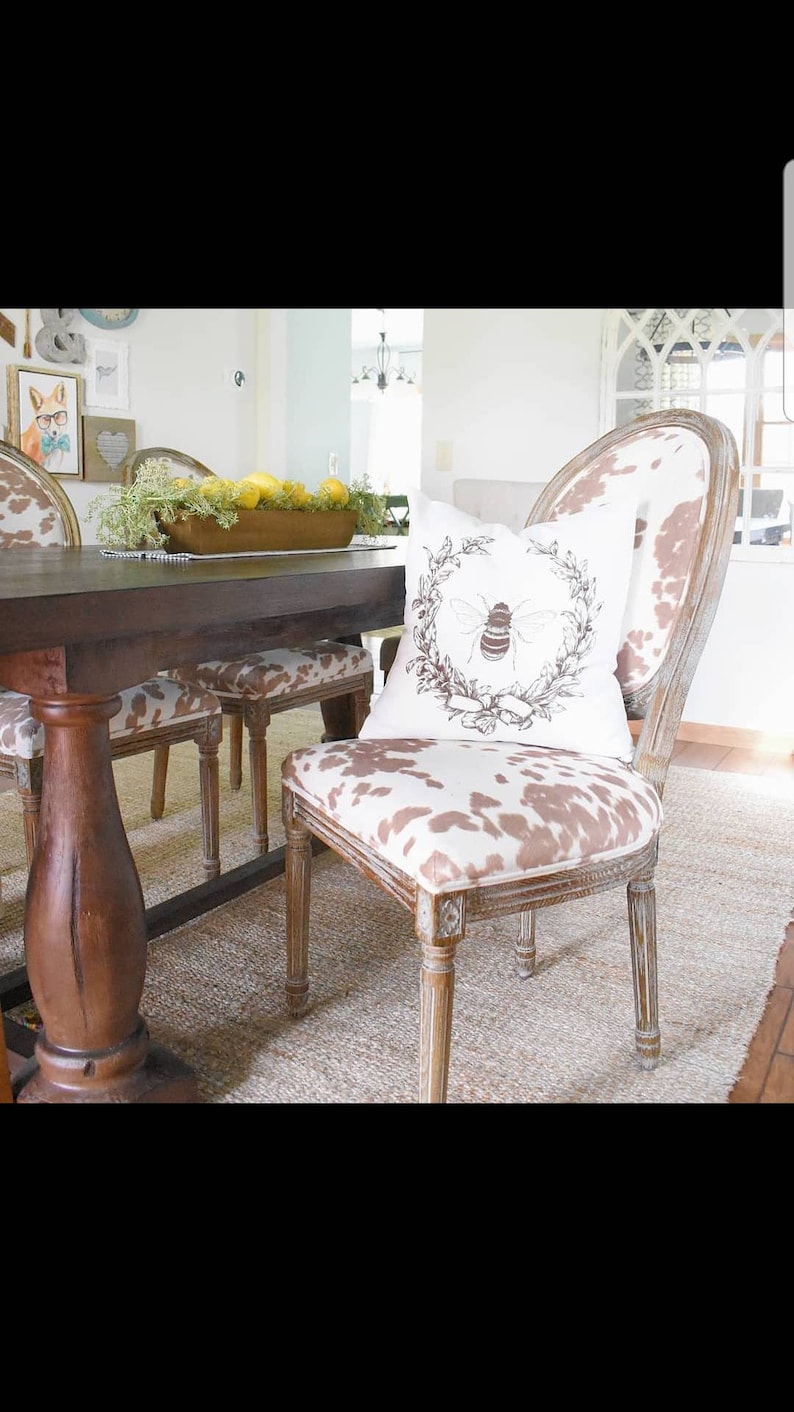 May include: A wooden dining table with a brown top and a single pedestal base.  The table is set with a bowl of lemons and greenery.  Two chairs with brown and white cowhide upholstery and a bee-themed pillow are visible.