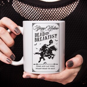 Op de afbeelding: Een witte keramische mok met een zwarte grafische print van een ruiter zonder hoofd op een paard. De tekst luidt Sleepy Hollow DEAD AND BREAKFAST en COME AND LAY YOUR WEARY HEAD TO REST, omringd door kleine vleermuizen en decoratieve randen.
