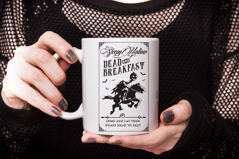 Op de afbeelding: Een witte keramische mok met een zwarte grafische print van een ruiter zonder hoofd op een paard. De tekst luidt Sleepy Hollow DEAD AND BREAKFAST en COME AND LAY YOUR WEARY HEAD TO REST, omringd door kleine vleermuizen en decoratieve randen.