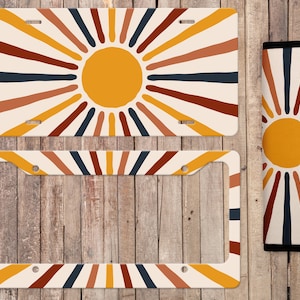 Puede incluir: Un juego de dos marcos para placas de matrícula con un diseño de rayos de sol y un sol amarillo en el centro. Los marcos son blancos con rayas rojas, naranjas y azules. El juego también incluye una cubierta para cinturón de seguridad de coche a juego con el diseño de rayos de sol.