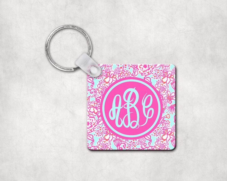 May include: A pink and blue floral patterned keychain with a silver key ring and a pink circle with a white monogram "ABC" in the center.