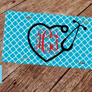 May include: A turquoise blue license plate with a white and turquoise pattern. A black heart with a red monogram and a black stethoscope are on the plate.