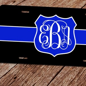 May include: Black license plate with a blue stripe and a blue and white monogrammed shield. The shield features the letters "EB" in a decorative font. The license plate has four mounting holes.
