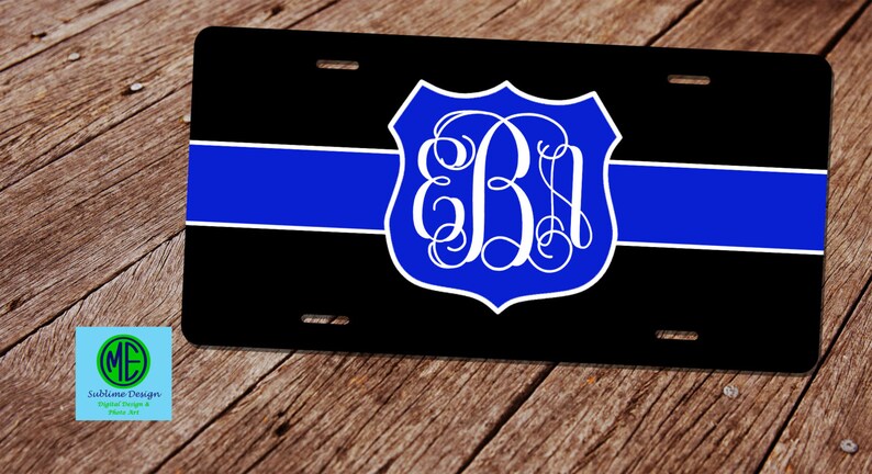 May include: Black license plate with a blue stripe and a blue and white monogrammed shield. The shield features the letters "EB" in a decorative font. The license plate has four mounting holes.
