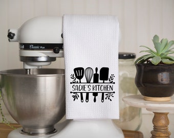 May include: White kitchen towel with black text that reads "Sadie's Kitchen" and a graphic of kitchen utensils. The towel is hanging on a white stand mixer.
