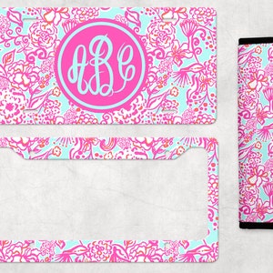 May include: A pink and blue floral patterned license plate frame with a monogrammed circle in the center. The monogram is "ABC" in white. The frame is surrounded by a white border. The image also includes a matching floral patterned license plate holder.