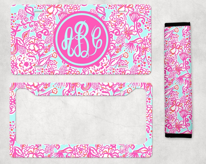 May include: A pink and blue floral patterned license plate frame with a monogrammed circle in the center. The monogram is "ABC" in white. The frame is surrounded by a white border. The image also includes a matching floral patterned license plate holder.