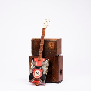 Puede incluir: Una guitarra roja, negra y blanca con un gráfico de un búfalo y el texto "FREMONT & JACKSON" en el cuerpo. La guitarra está sentada encima de dos cajas de madera, una encima de la otra.