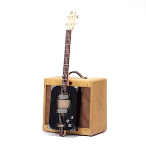 Puede incluir: Una guitarra eléctrica negra con un mástil de madera y una sola pastilla está montada en un amplificador beige de estilo vintage.