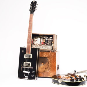 May include: Two electric guitars and a vintage amplifier. One guitar is black with a hollow body and the other is brown with a solid body.