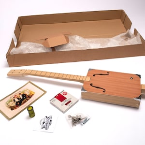 Puede incluir: Un kit de guitarra de caja de puros con un cuerpo de madera, un mástil de madera y un juego de cuerdas. El kit también incluye una clavija de afinación, un puente y un juego de tornillos. El kit está empaquetado en una caja de cartón marrón.