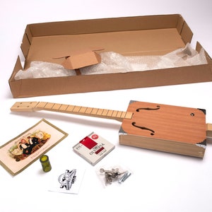 Puede incluir: Un kit de guitarra de caja de puros con un cuerpo de madera, un mástil de madera y un juego de cuerdas. El kit también incluye una clavija de afinación, un puente y un juego de tornillos. El kit está empaquetado en una caja de cartón marrón.