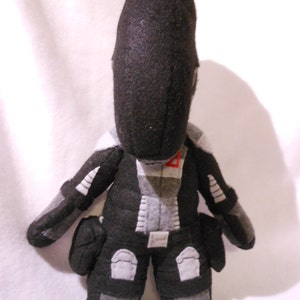 May include: Black and grey plush toy with a humanoid shape and a large head. The toy has a red detail on the chest and is wearing a black and grey suit with white accents.