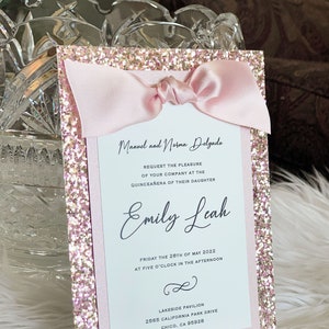 May include: Pink glitter card with a pink ribbon bow and white text that reads "Manuel and Norma Delgado request the pleasure of your company at the Quinceañera of their daughter Emily Leah Friday the 28th of May 2022 at five o'clock in the afternoon Lakeside Pavilion 2565 California Park Drive Chico, CA 95928"