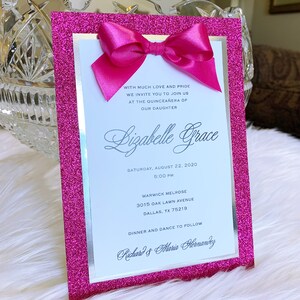 May include: Pink glitter card with a silver border and a pink bow. The card is an invitation to a Quincea&ntilde;era for Lizabelle Grace. The invitation includes the date, time, location, and the names of the hosts, Richard and Maria Hernandez.