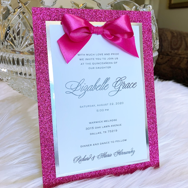 May include: Pink glitter card with a silver border and a pink bow. The card is an invitation to a Quincea&ntilde;era for Lizabelle Grace. The invitation includes the date, time, location, and the names of the hosts, Richard and Maria Hernandez.